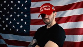 Patriotic white man with tattoos and beard putting on a red MAGA or Make America Great Again hat in slow motion in front of a waving american flag and looks directly at the camera with confidence. - Powered by Shutterstock - Get 15% off with code: PIKWIZARD15