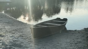 A small, empty wooden rowboat rests on the sandy shore of a calm lake, with golden sunlight reflecting on the water at sunrise or sunset - Powered by Shutterstock - Get 15% off with code: PIKWIZARD15