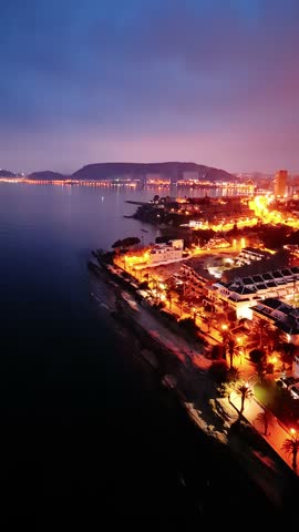Aerial footage of coastal city at night with streetlights, buildings, coastline, and reflections on water. Captured by drone during twilight.