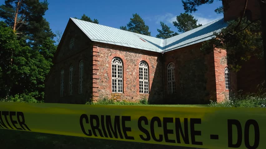 Police Crime Scene tape Do Not Enter in front of a large and old church that is made out of red bricks during a sunny day in a rural area with no people around. Yellow tape is moving in the wind slow.