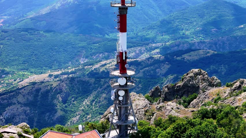 Cinematic aerial footage of a TV broadcasting tower above the city of Sliven, Bulgaria, filmed with a drone using a 77mm lens. The drone smoothly circles the tower, revealing dramatic rocky mountain 