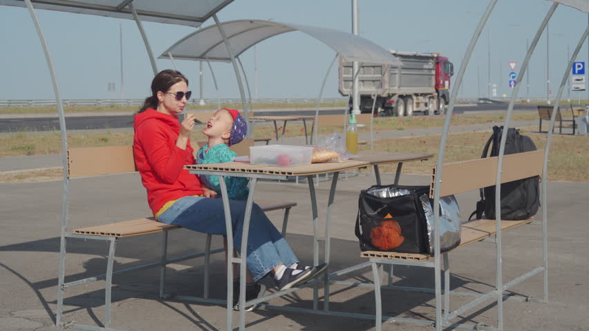 A person in a red top is seated at an outdoor table, while a child sits next to them, both enjoying a break. They are at a roadside rest area in Crimea under a sunny sky