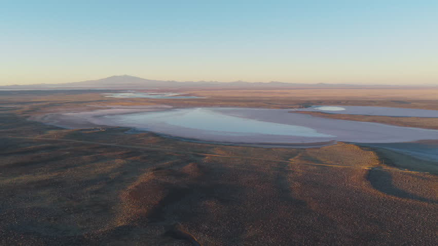 Aerial drone view of Salinas del Diamante basin in Mendoza, Argentina, reveals vast salt flats surrounded by arid terrain with the Andes in the distance under warm evening light