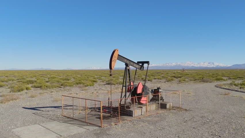 Drone captures oil pumpjack operating in dry steppe near Mendoza, Argentina, with snowy Andes peaks in background under clear blue sky, wide and slow pull away shot