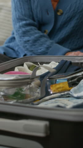 Vertical shot of elderly woman sitting in front of camera and folding necktie of her husband, then putting it into suitcase with other items for traveling while packing baggage