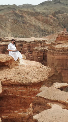 Woman in white dress working on laptop while sitting on canyon cliff, digital nomad and remote work lifestyle concept in desert landscape outdoors.