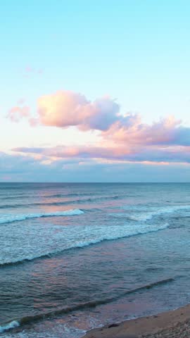 Cloudy, bright blue sky over a grey beach. Waves crash against the shoreline, scattering pebbles. It�s a peaceful, natural coastal scene.