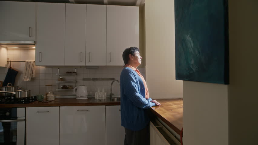 Senior man walking up to his wife standing by window in domestic kitchen, touching her shoulders and embracing woman while both expressing affection and love