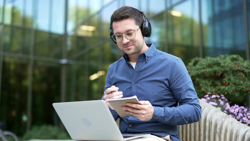 Businessman in headphones watching video call online conference notes in notebook sitting outside on a bench near office building. Worker communicates remotely at business meetings, training. Close up