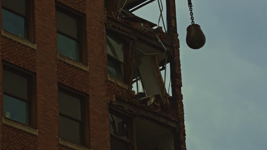 Close-up shot of a wrecking ball smashing through the upper floors of a partially demolished brick high-rise during a downtown demolition project.