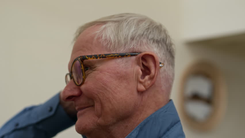 Tilt down of modern elderly man in eyeglasses buttoning collar of new blue shirt and touching his grey hair while getting dressed in front of mirror in corridor