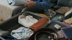 Elderly woman in blue knitted cardigan sitting on sofa in home environment and putting folded things into suitcase for baggage while preparing for travel - Powered by Shutterstock - Get 15% off with code: PIKWIZARD15