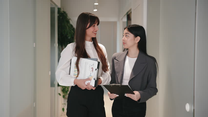 Positive Asian female with straight hair smiling and posing beside confident coworker holding tablet. Standing in hallway while preparing for presentation or welcoming business partners. - Powered by Shutterstock - Get 15% off with code: PIKWIZARD15
