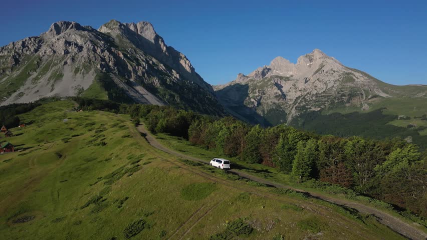 White car driving along picturesque mountain ridge in the morning through green meadows crossing idyllic alpine landscape with high mountains in the background, aerial view