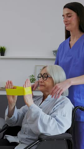 Vertical HD video of mid adult female nurse helping a senior woman in a wheelchair doing exercises with resistance band for rehabilitation at home
