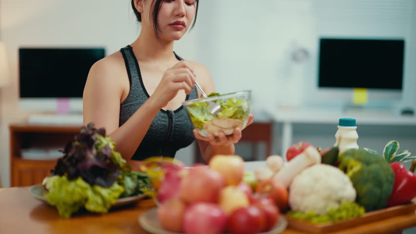 Young asian sportswoman sitting at table with fresh vegetables and fruits, showing clear disgust while attempting to eat salad, expressing reluctance toward healthy nutrition and diet challenges
