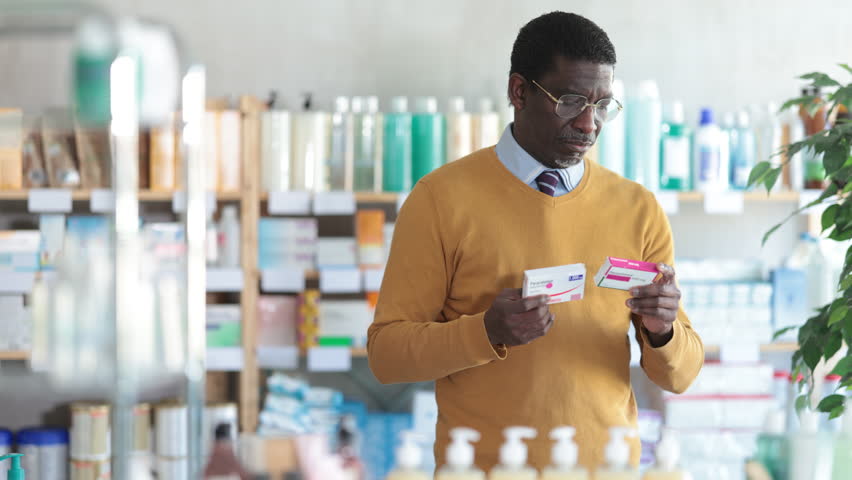 African man choose buy painkiller paracetamol tablet at pharmacy, compare with other good. Client check for absence of harmful substances, view product in details.