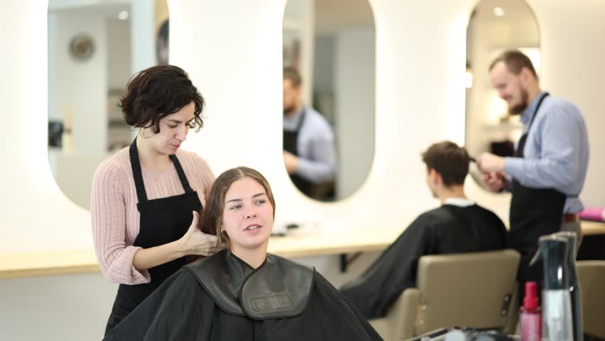 Young female hairdresser stylist gives haircut to adult female client in salon