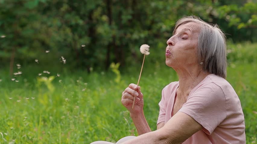 An elderly woman in a park, smiling and enjoying the moment as she holds a dandelion and watches the seeds float away.