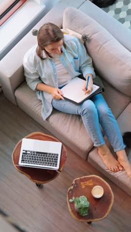 Overhead view of woman writing notes, lying on couch at home