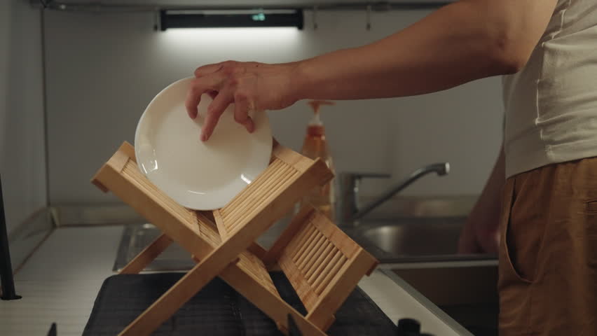Close-up cropped shot of unrecognizable man placing clean white dishes and drinking glass on wooden drying rack in modern kitchen, slow motion. Concept of tidiness and organization after dishwashing.