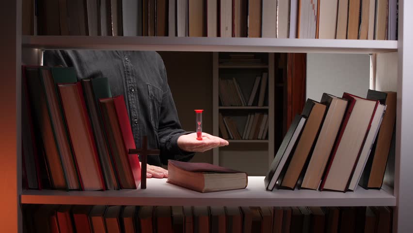 priest holding hourglass in library. Concept of transience of time, religion, learning