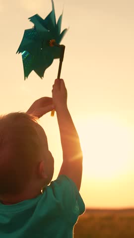 Kid playing with toy pinwheel in hand on summer field, sunny day. Childhood of boy. Happy child having fun with toy spinner outside in summer park. Family vacation, nature. Children outdoors. Baby sun