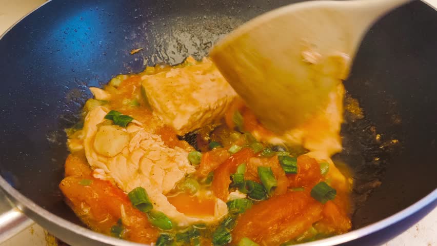 Close-up view of cooking hot, flavorful salmon with tomato sauce and sliced green onions in a sizzling frying pan on high heat. Indoor kitchen scene showcasing a delicious, freshly prepared meal.