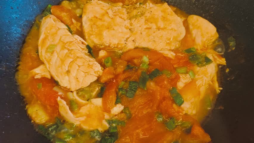 Close-up view of cooking hot, flavorful salmon with tomato sauce and sliced green onions in a sizzling frying pan on high heat. Indoor kitchen scene showcasing a delicious, freshly prepared meal.