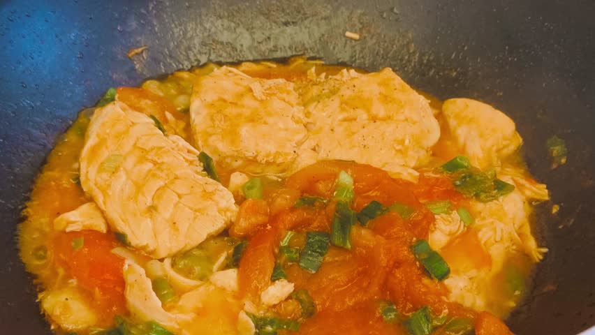 Close-up view of cooking hot, flavorful salmon with tomato sauce and sliced green onions in a sizzling frying pan on high heat. Indoor kitchen scene showcasing a delicious, freshly prepared meal.