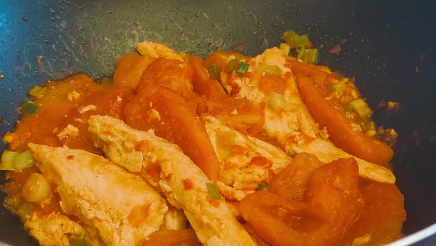Close-up view of cooking hot, flavorful salmon with tomato sauce and sliced green onions in a sizzling frying pan on high heat. Indoor kitchen scene showcasing a delicious, freshly prepared meal.