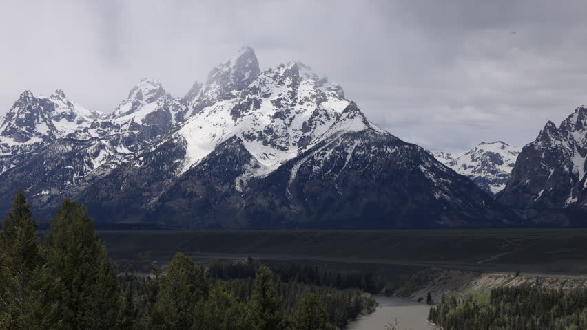 Snow-capped Grand Teton and the Cathedral Group shine under a vibrant blue sky, framed by evergreens and drifting clouds, as seen from Snake River Overlook in Wyoming