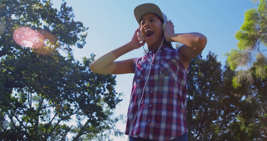 Woman holding headphones while singing to music in sunlit park, generating blue swirls around torso. Music, rhythm, vibrancy, expression, outdoor, serenity, movement