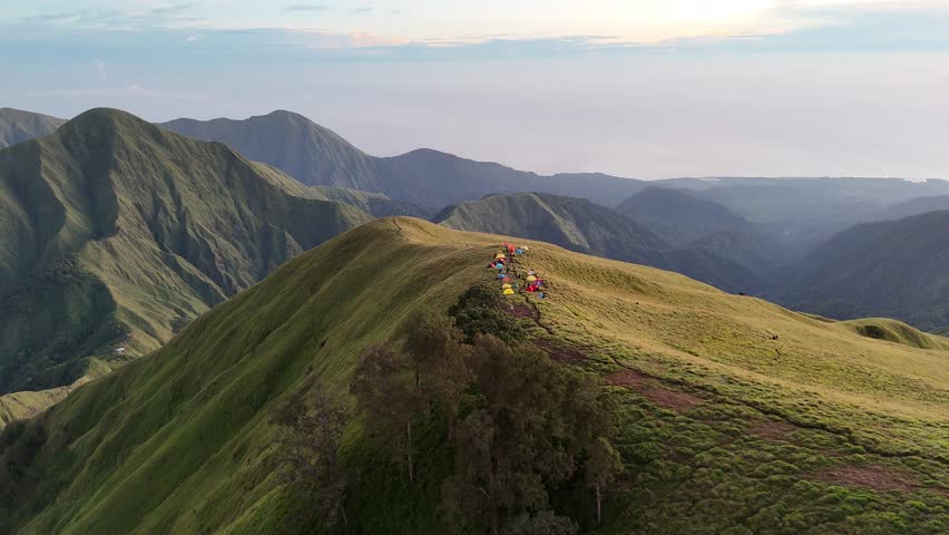 A stunning, static landscape view of the Mount Rinjani volcano from the crater rim campsite, with colorful hiker tents in the foreground under a clear blue sky on Lombok, Indonesia
