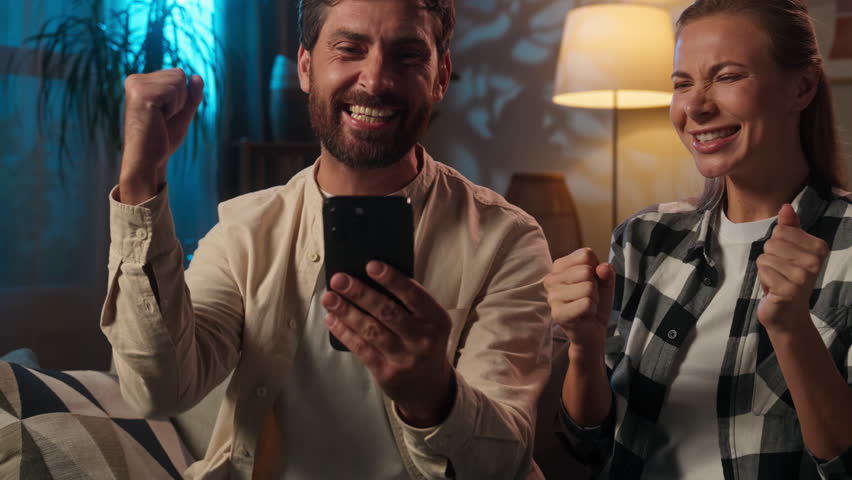 Caucasian couple happy woman man guy girl cheerful husband wife family wish achieve smartphone victory celebrating mobile phone win rejoice scream cheer online gambling betting prize at evening home
