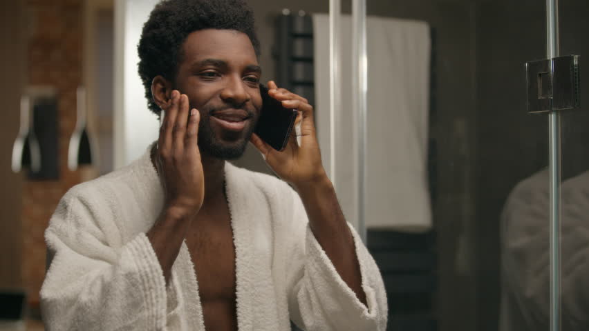 African American man in bathroom talking mobile phone guy after shower morning shaving beard touching smiling bearded male call friendly conversation speak cellphone communication in hotel bath home