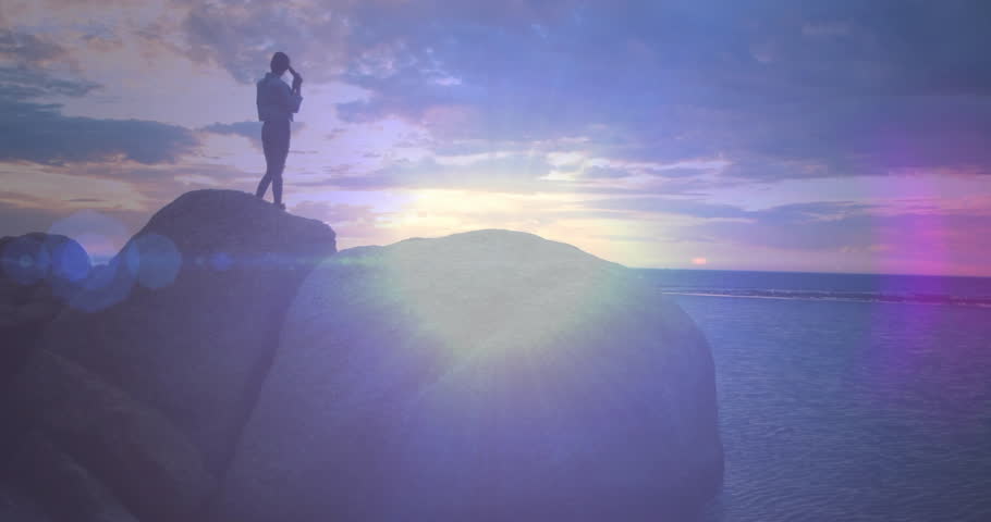 Traveler climbing boulder at sunset, sun flaring shielding eyes holding smartphone gazing at sea. Adventure, exploration, nature, rugged, tranquility, outdoor, wilderness