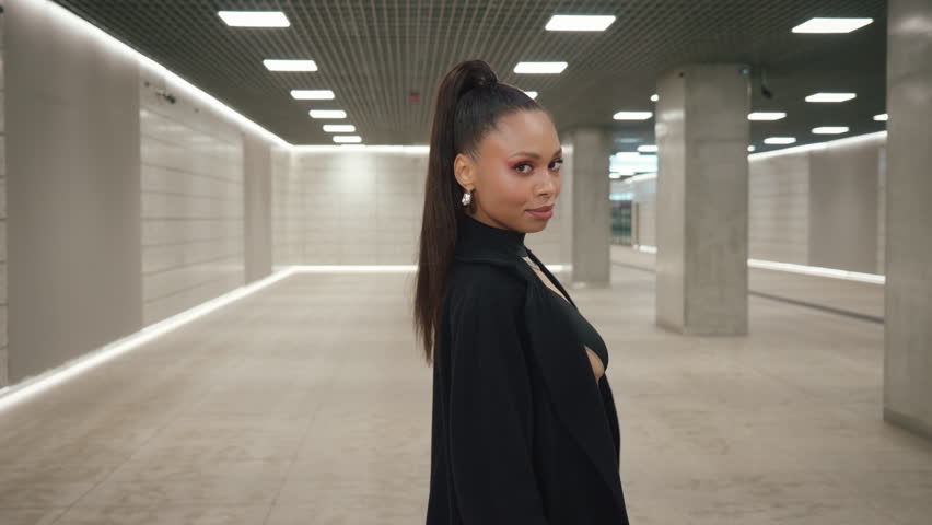 Following shot portrait of happy Latina female walking through modern station tunnel hall. Brunette woman wearing long black coat walks looking at camera and smiling. Futuristic architecture