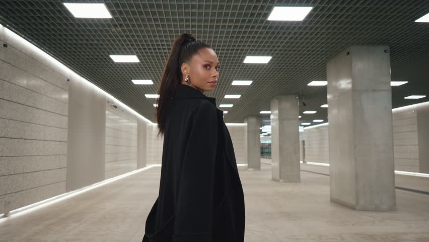 Following shot portrait of happy Latina female walking through modern station tunnel hall. Brunette woman wearing long black coat walks looking at camera and smiling. Futuristic architecture