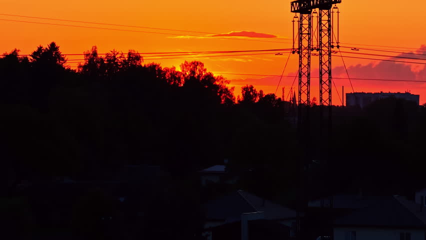 Silhouette of a large power line pylon against a stunning, fiery orange sunset over a forested landscape in Latvia, blending nature and industry - An aerial drone shot
