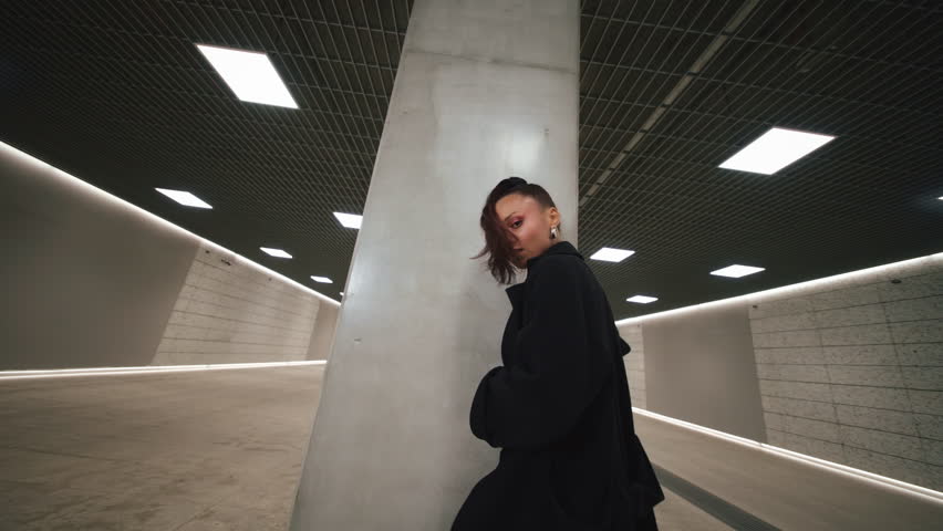 Confident Latina woman dancing seductively and looking at camera in futuristic underground subway hall tunnel. Sexy dance, female in black bodysuit, thigh-high boots and coat, concrete walls