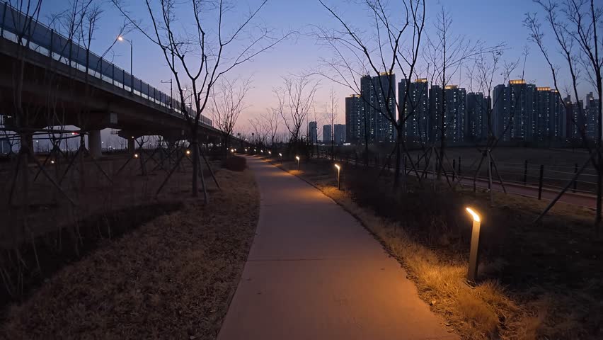 Lit Paved Pathway At Dusk Alongside Road And Bridge in Incheon, South Korea. City Buildings In Background. wide pullback shot