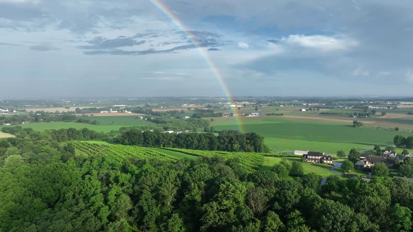 Aerial view of rural Pennsylvania, showing lush green fields and rolling hills under vast sky. Vibrant rainbow arches over landscape, adding touch of magic to serene countryside scene. Wide shot.