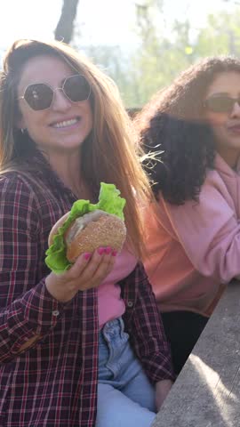Two female friends enjoying burgers while sitting at wooden picnic table, sharing laughter and delicious meal during sunny summer day in park