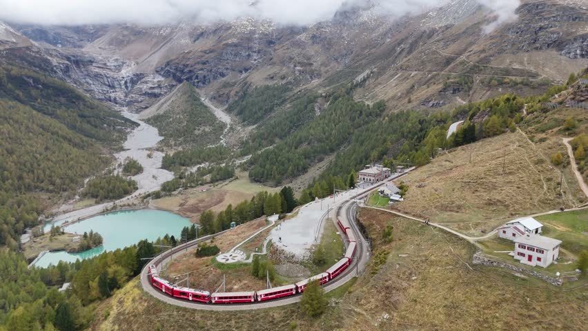 A train of Rhaetian Railway snakes thru the famous curve near Alp Grüm Station on the Bernina Line, in Poschiavo, Graubünden, Switzerland, overlooking Lake Palu below the receding glacier in bacground