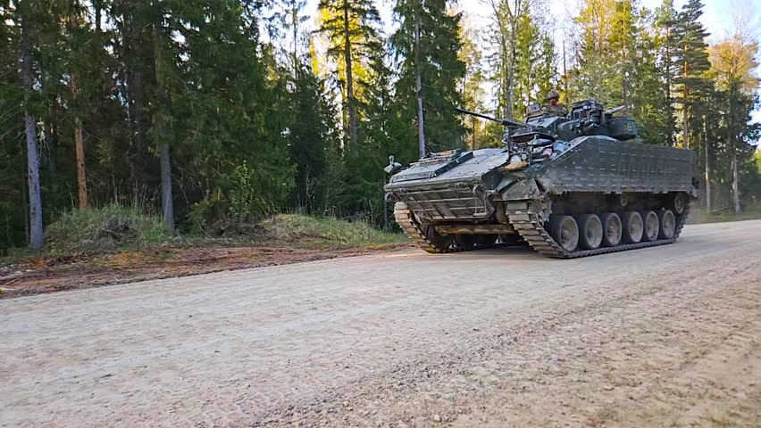 Gorgeous footage of a British Warrior FV510 armoured personnel carrier tank driving on a gravel road in a rural countryside sunny forest with a dust cloud behind it and soldiers with weapons on top.