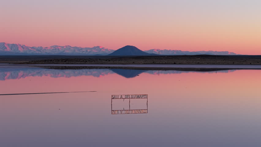 Low drone pullback over calm reflective waters of Salinas del Diamante at sunset, showing the conical silhouette of Volcán and the snowcapped Andes in Mendoza, Argentina