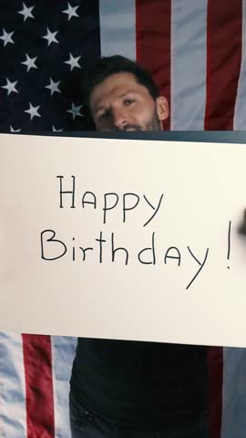 Tattooed white man lifts up a white notebook that has Happy Birthday written in it in front of American vertical flag showing he celebrates 4th of July American Independence day with happines and joy.