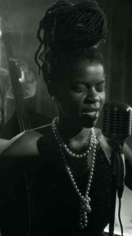 Vertical black and white shot of glamorous Black woman singing to microphone and swaying to music while performing on stage in classic retro style club