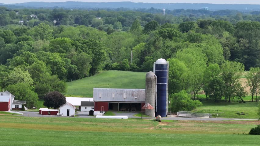 Aerial zoom shot of american farmstead with red barn, silo storage and house in front of green forest landscape. Dairy and fields in rural area. Pennsylvania, USA. Panorama